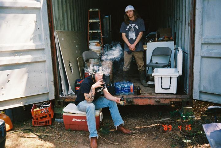 Two men stand and sit in the open back of a storage container, one smoking a cigar and the other looking at the camera.