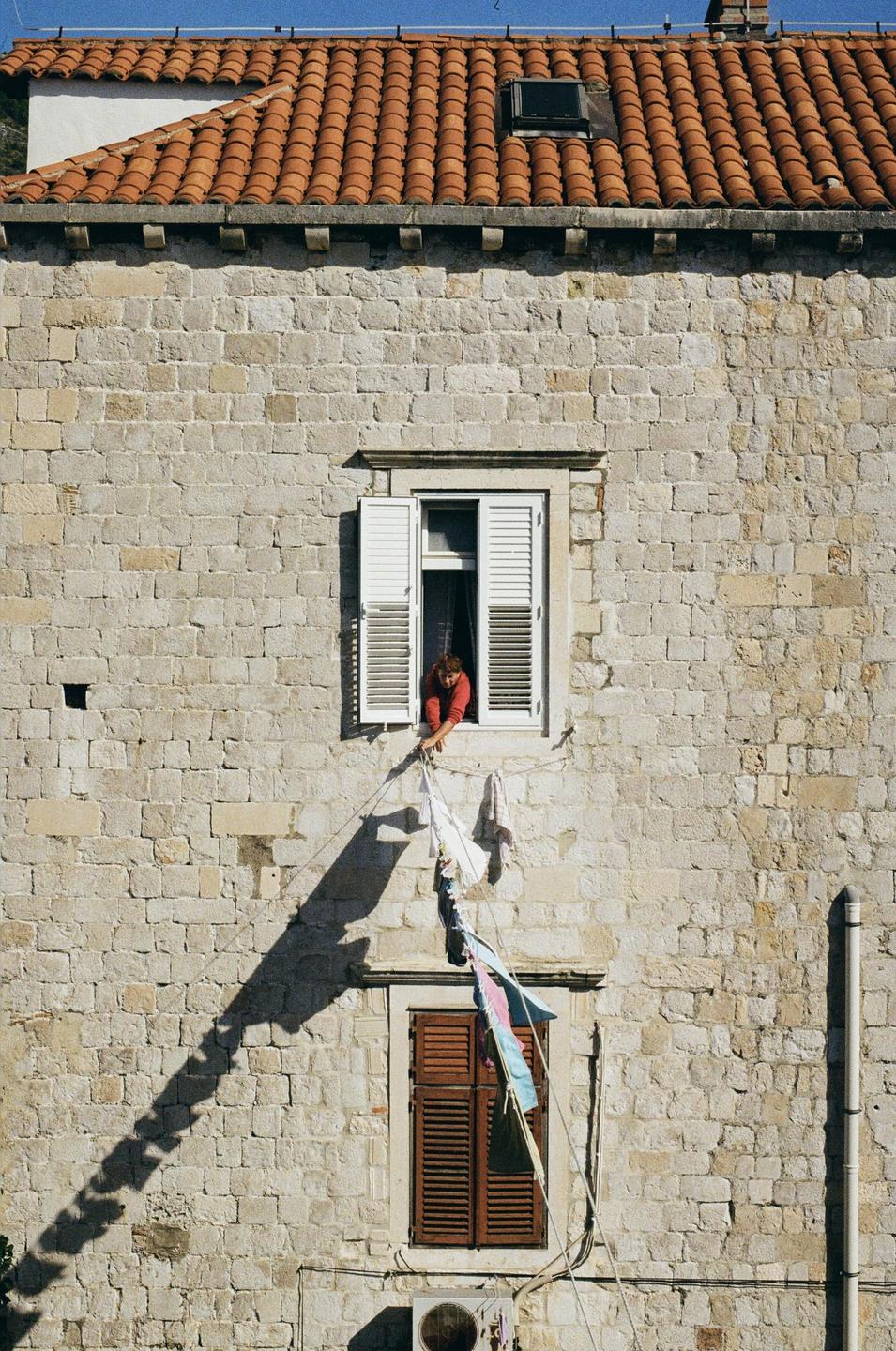 A woman leans out of a window with white shutters to hang laundry on a clothesline.