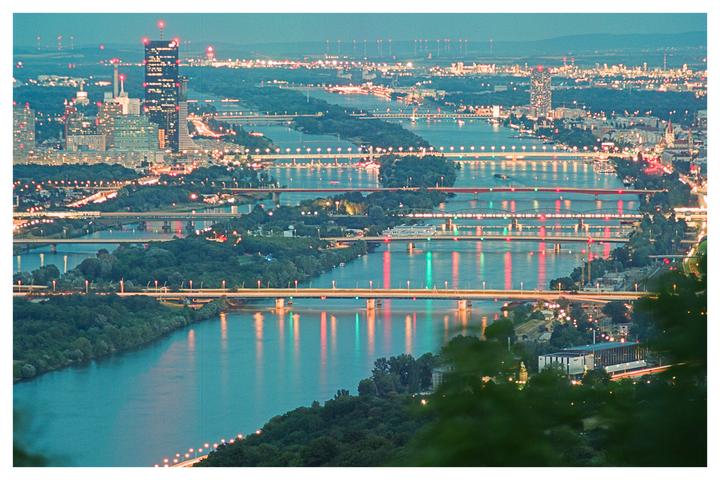 A high-angle view shows the city of Vienna at twilight, with multiple bridges crossing a river and city lights reflecting on the water.