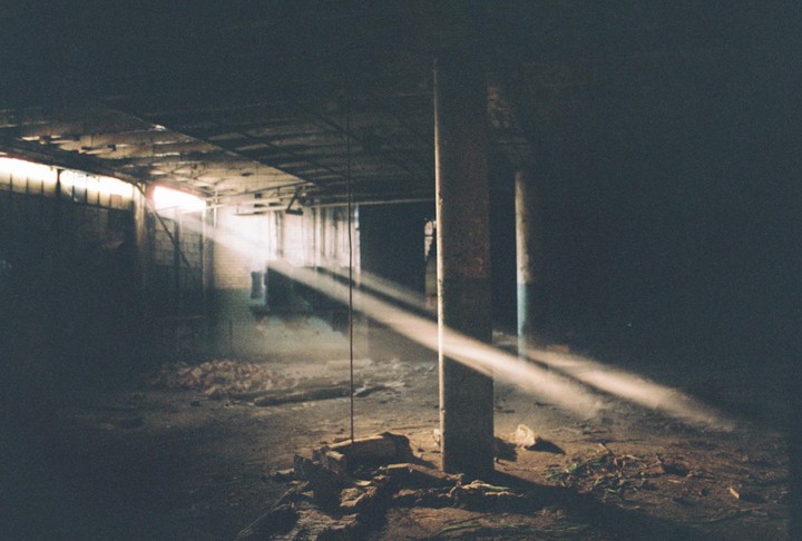 Sunlight streams through a window into a dusty, abandoned industrial building with concrete pillars and debris on the floor.