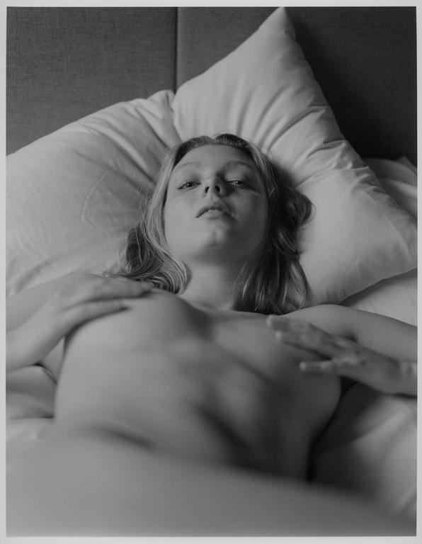 A woman lies topless on a bed with her hands covering her chest.