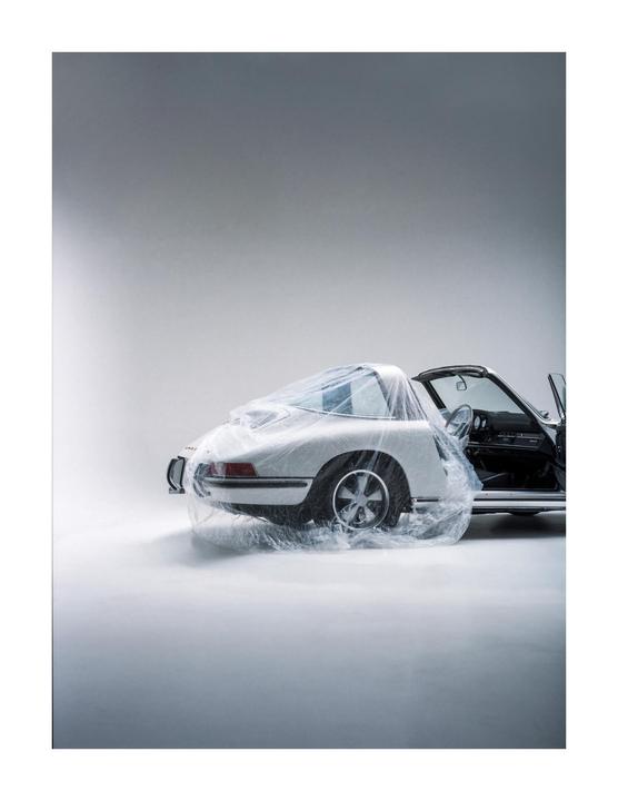 A white vintage car is partially covered in clear plastic sheeting with its door open.