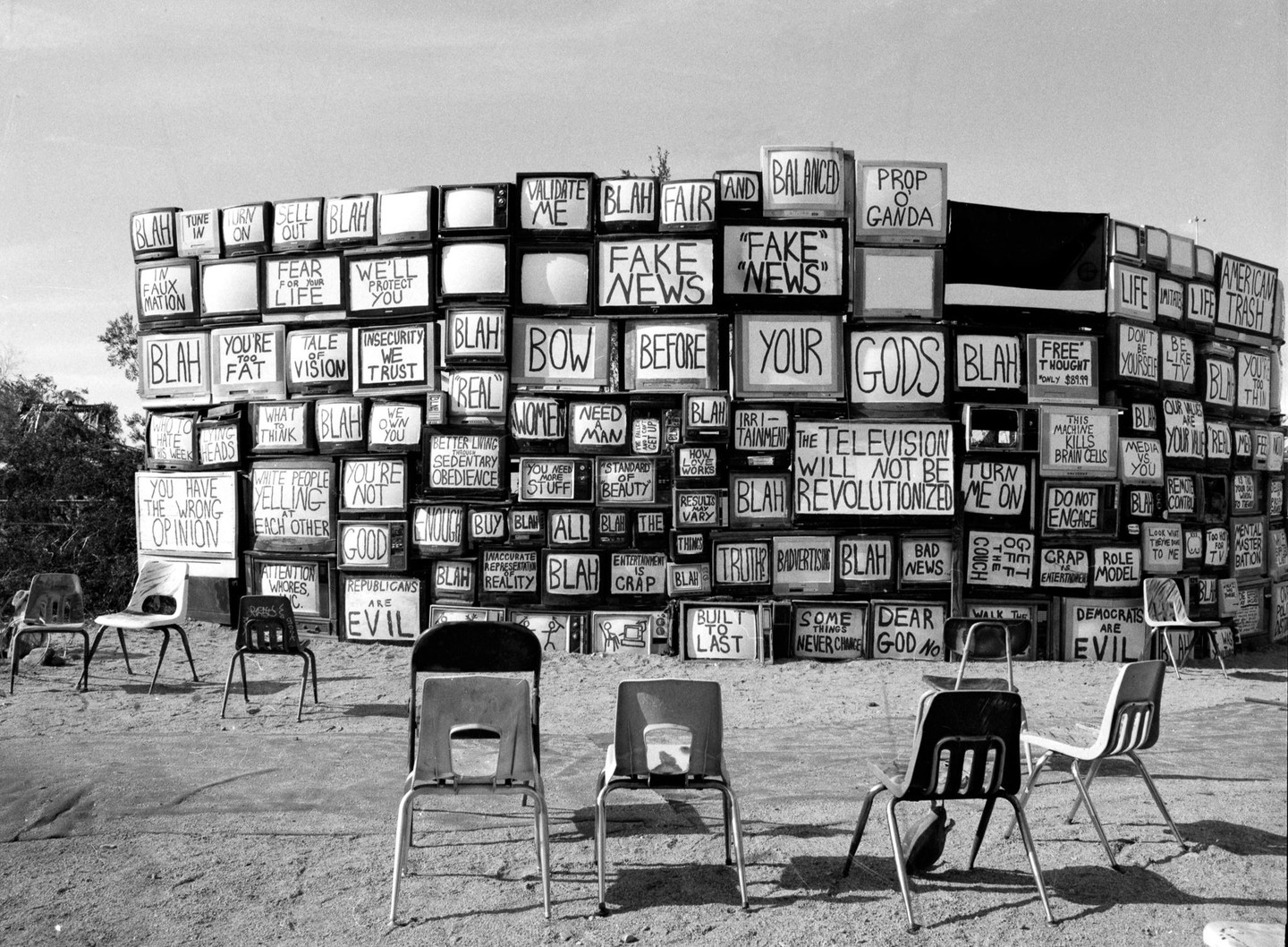 A wall of old television sets with various phrases painted on their screens stands in a desert-like setting, with several chairs arranged in front of it.