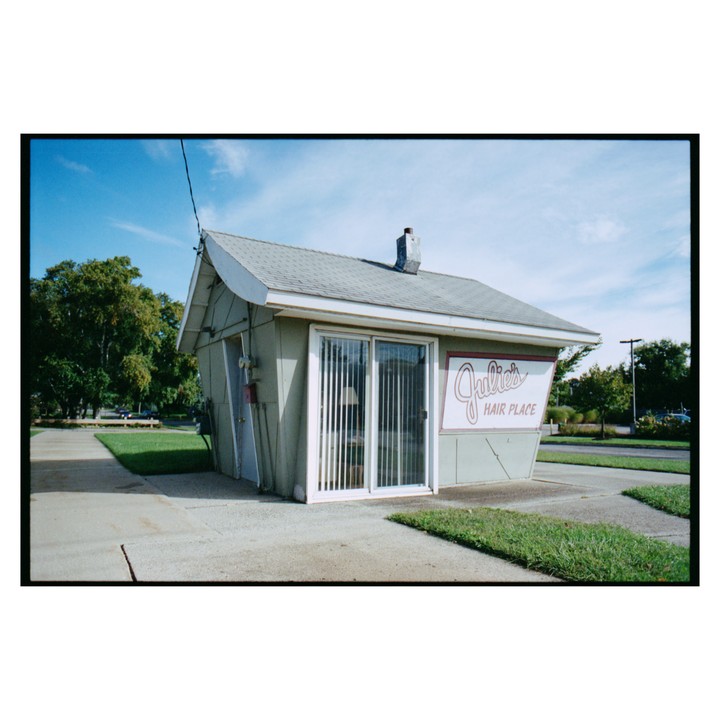 A small, light green building with a sign that reads "Julie's Hair Place" stands next to a sidewalk and grass.