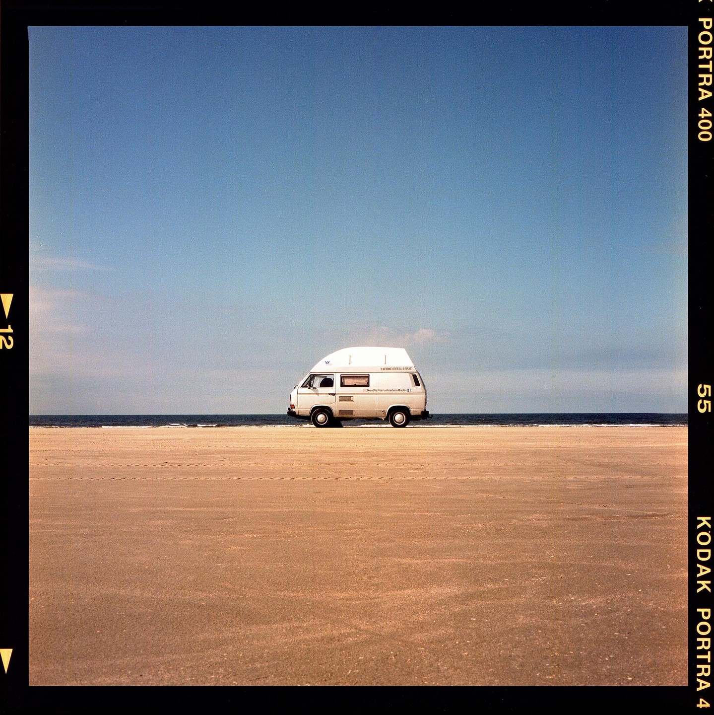 A white camper van is parked on a sandy beach next to the ocean under a clear blue sky.