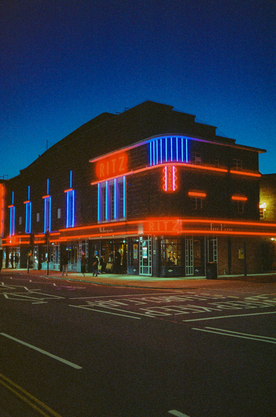 A building with neon signs for "Ritz" is illuminated at night, with a street and people visible below.