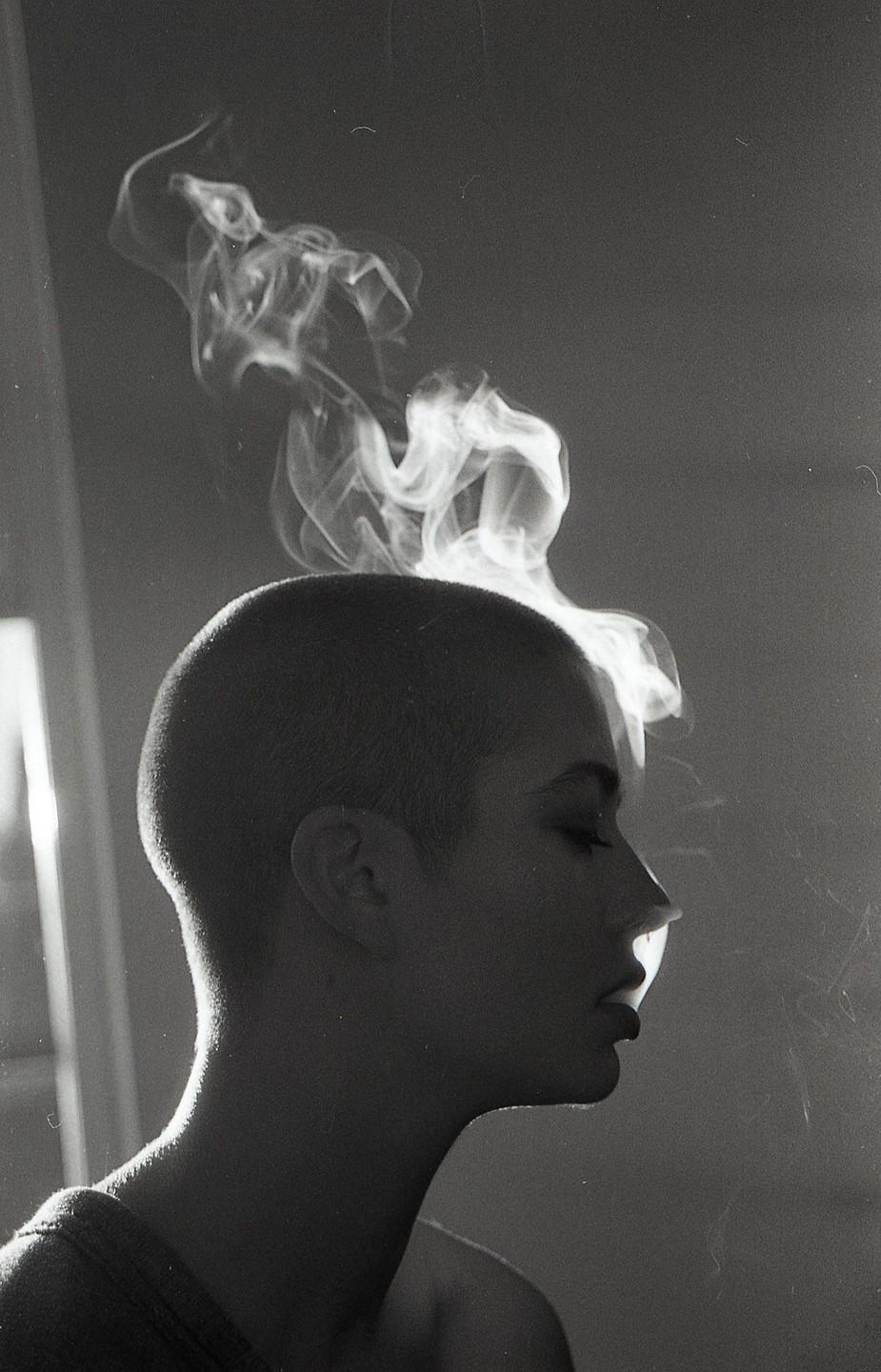 A woman with a shaved head is seen in profile, exhaling smoke that billows above her head.