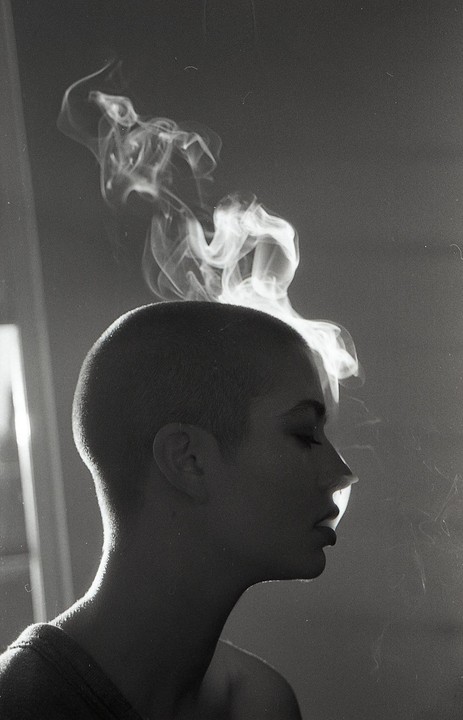 A woman with a shaved head is seen in profile, exhaling smoke that billows above her head.