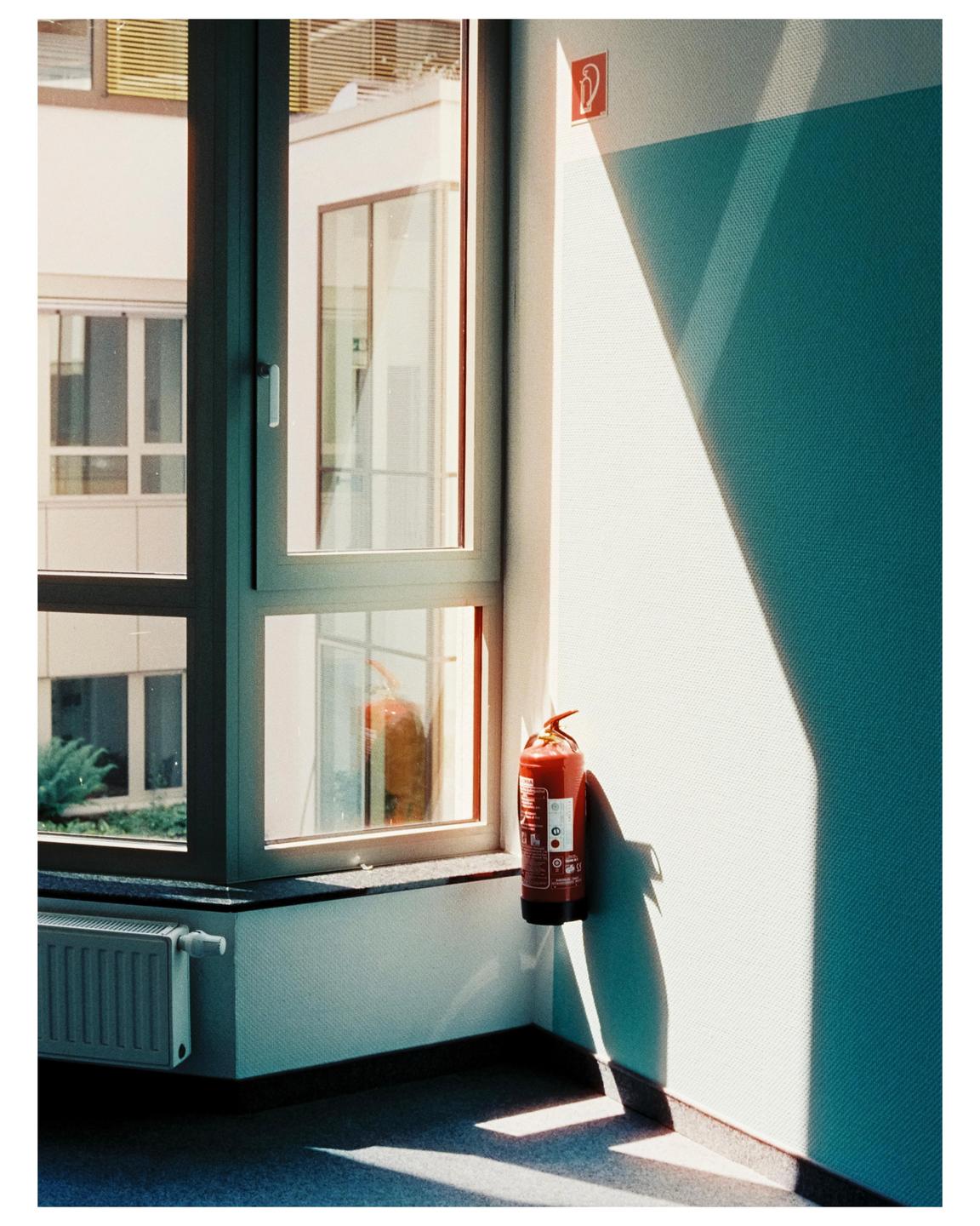A fire extinguisher hangs on a wall next to a window, with sunlight casting shadows on the wall and floor.