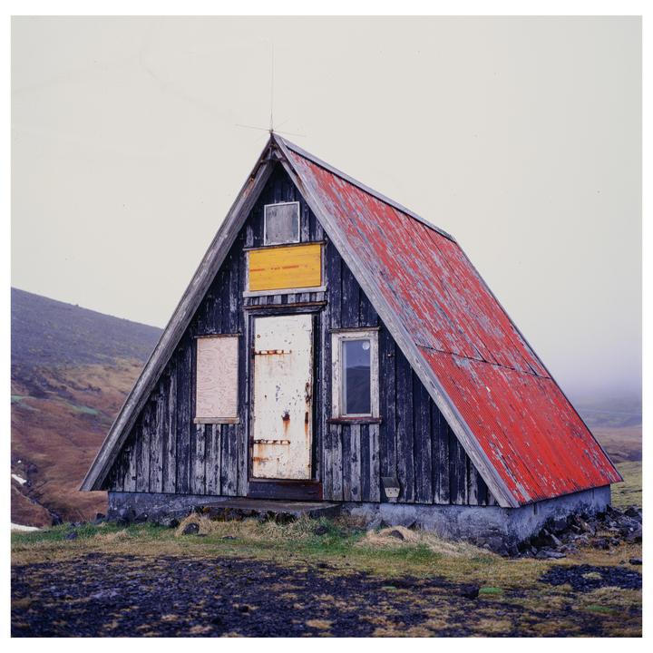 An A-frame cabin with a red roof stands on a grassy hill under a white sky.