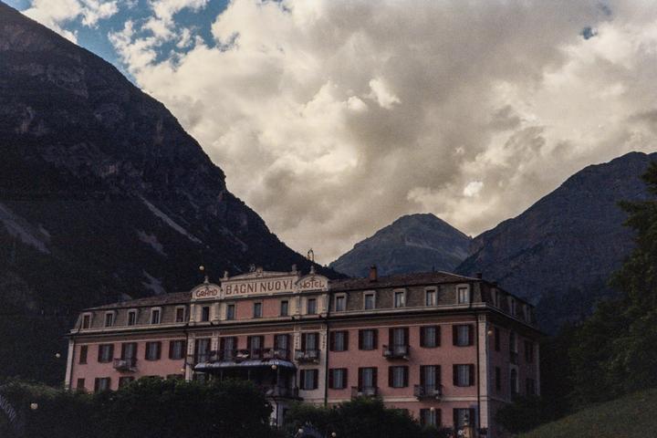 A large pink hotel with many windows and balconies sits at the base of a mountain range under a cloudy sky.