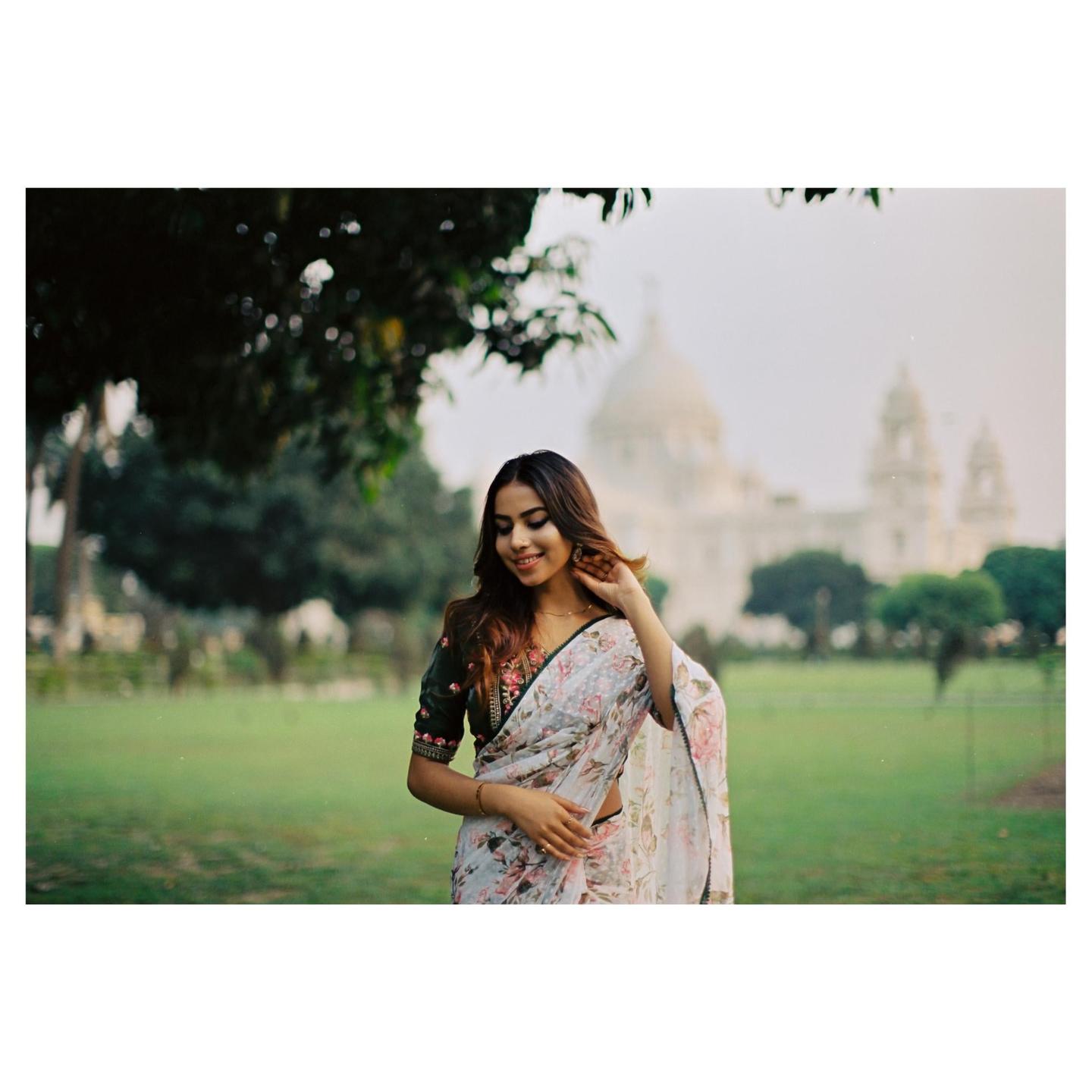 A woman in a floral sari stands in a grassy park with a large domed building in the background.