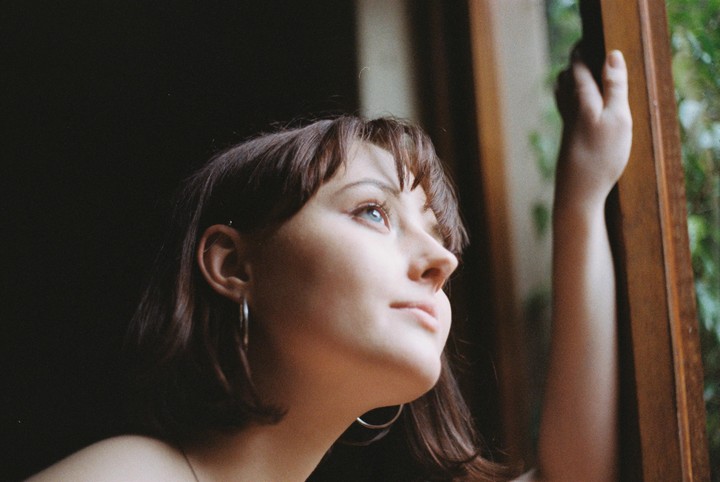 A woman with bangs and hoop earrings looks up and out a window, holding the wooden frame.