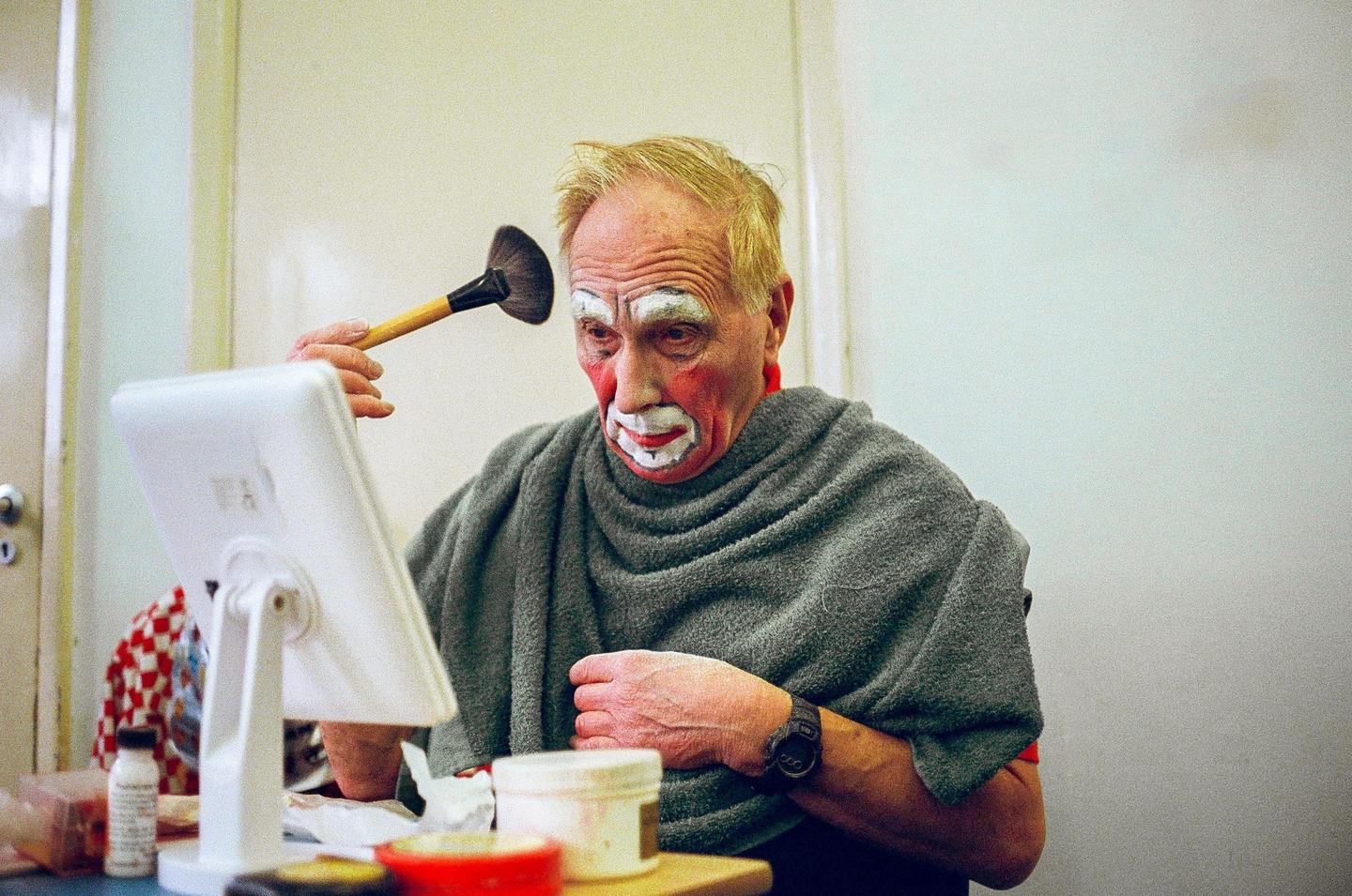 A man with clown makeup on his face applies more makeup with a brush while looking in a mirror.