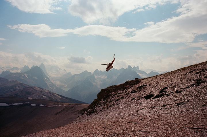 A red and white helicopter flies over a rocky mountain slope with a range of mountains in the background under a cloudy sky.