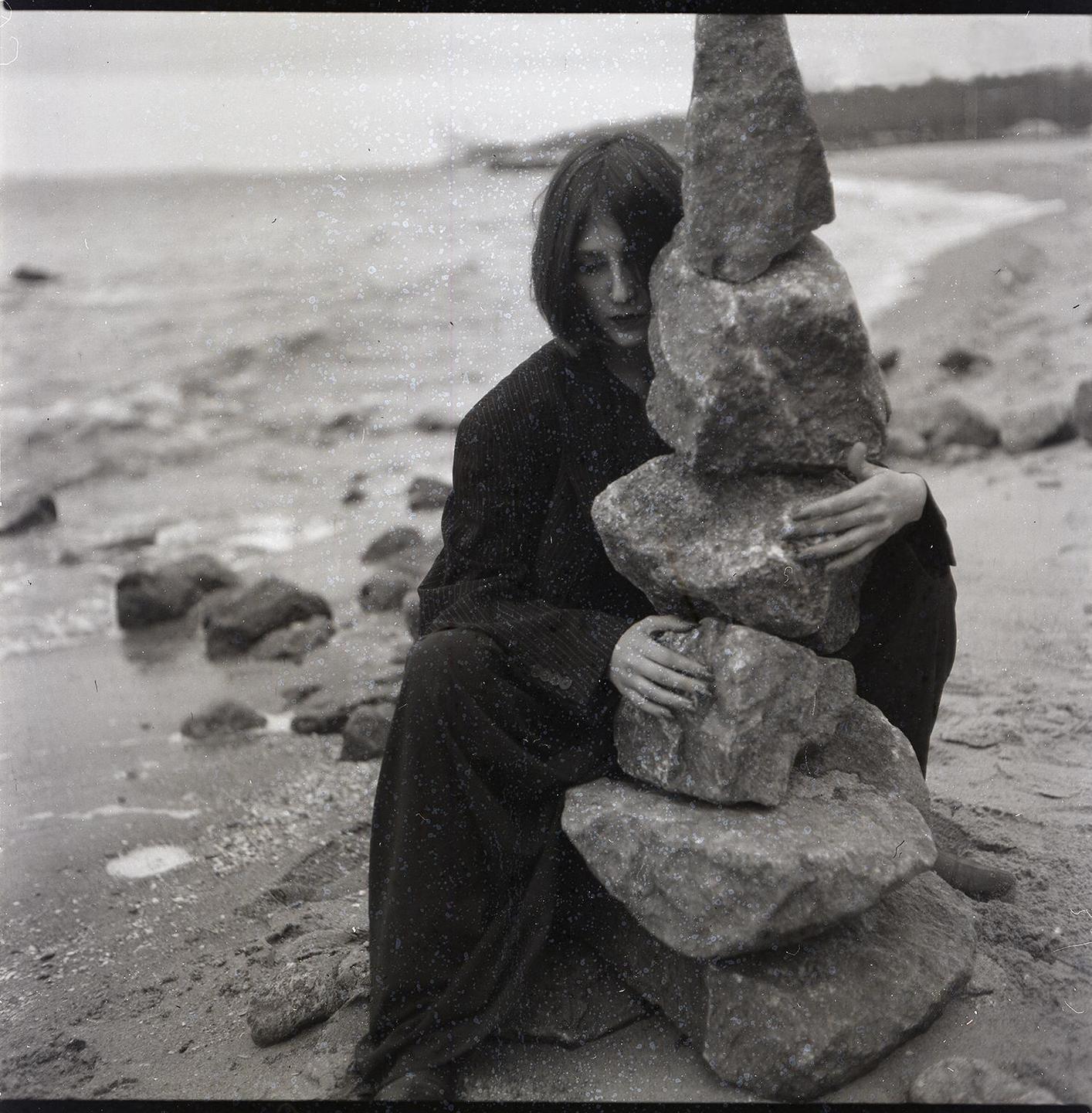 A woman sits on a rocky beach, holding a stack of balanced rocks.