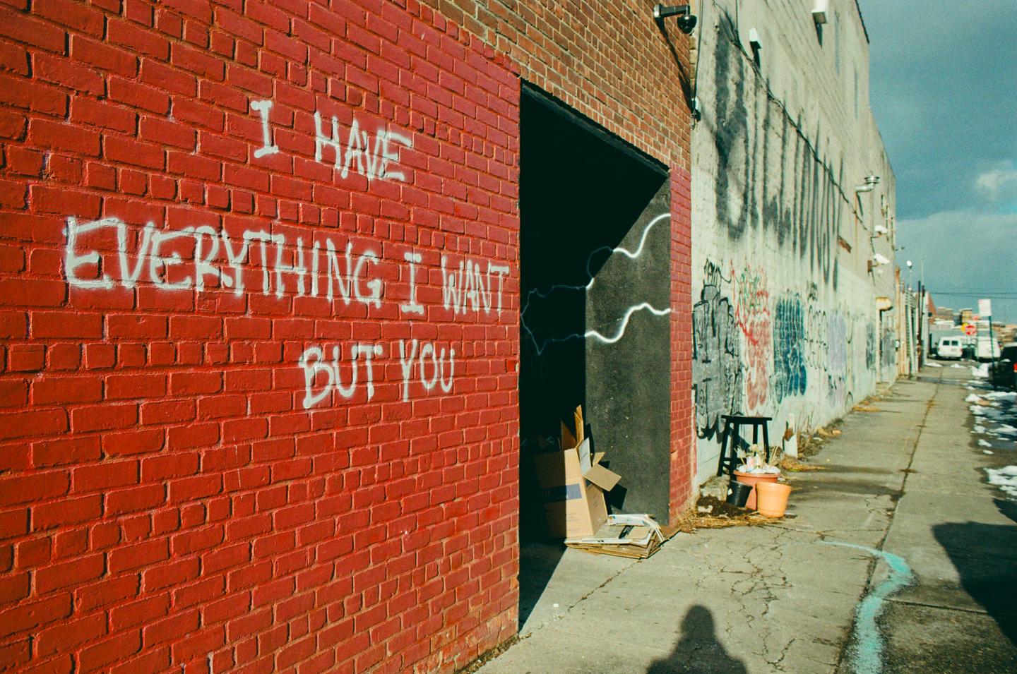 White graffiti on a red brick wall reads "I HAVE EVERYTHING I WANT BUT YOU" next to an alleyway with more graffiti and a shadow of a person.