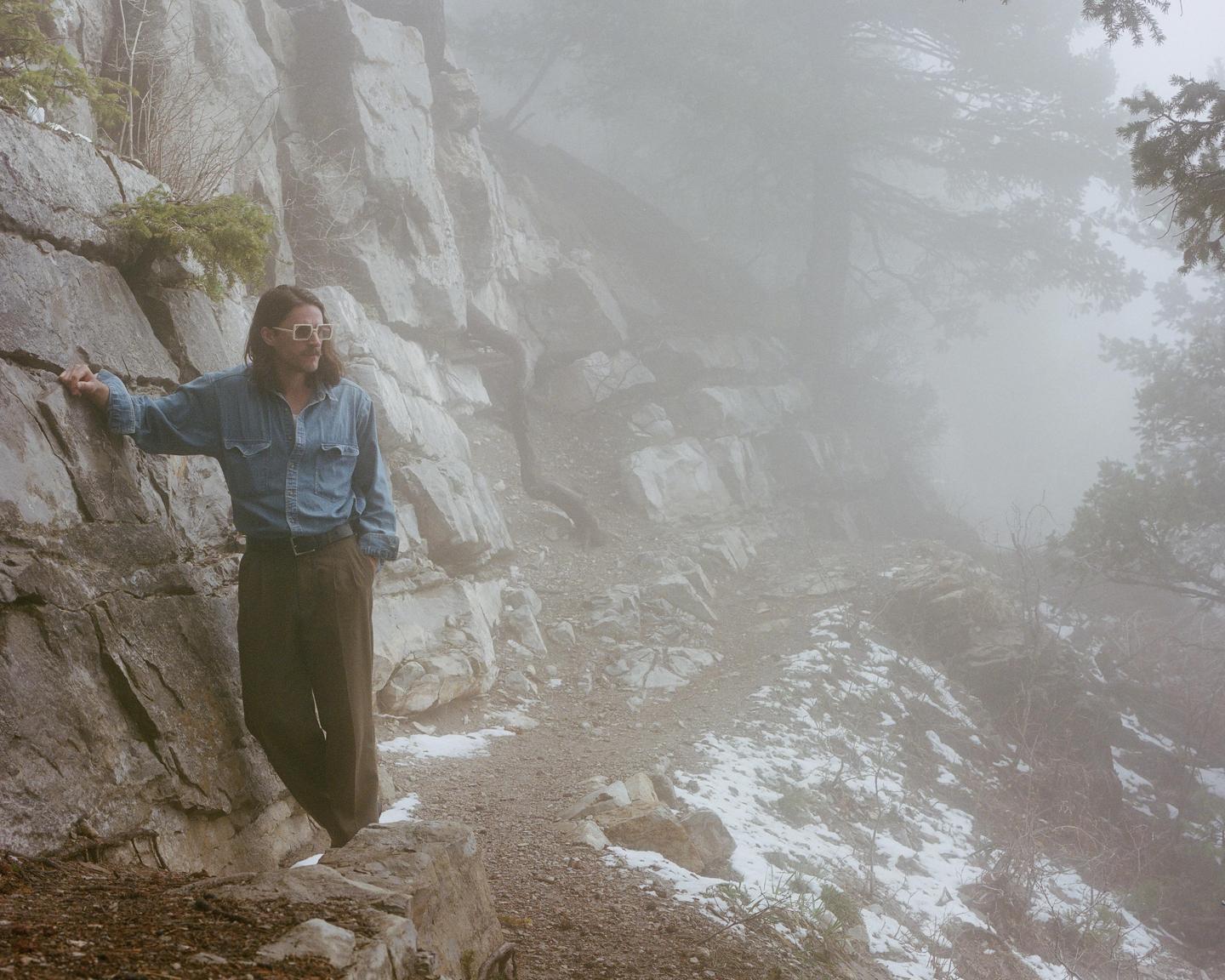 A man stands on a snowy mountain path next to a rock face in the fog.