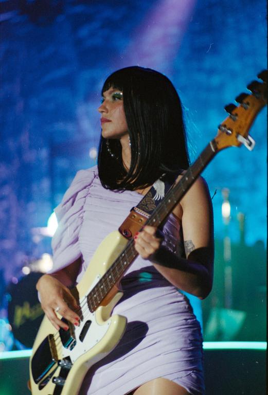 A woman with dark hair and a purple dress plays a bass guitar on a stage with blue lighting.