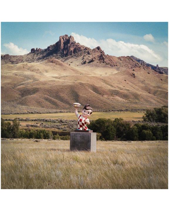 A statue of a boy holding a hamburger stands on a concrete pedestal in a grassy field with mountains in the background.