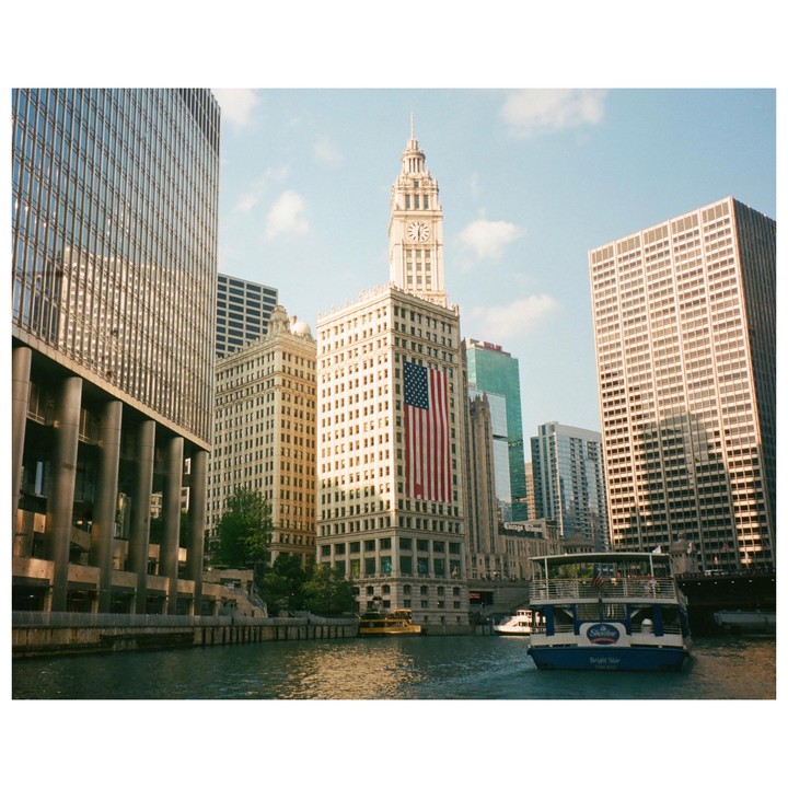 A boat travels on a river surrounded by tall buildings, one of which displays a large American flag.