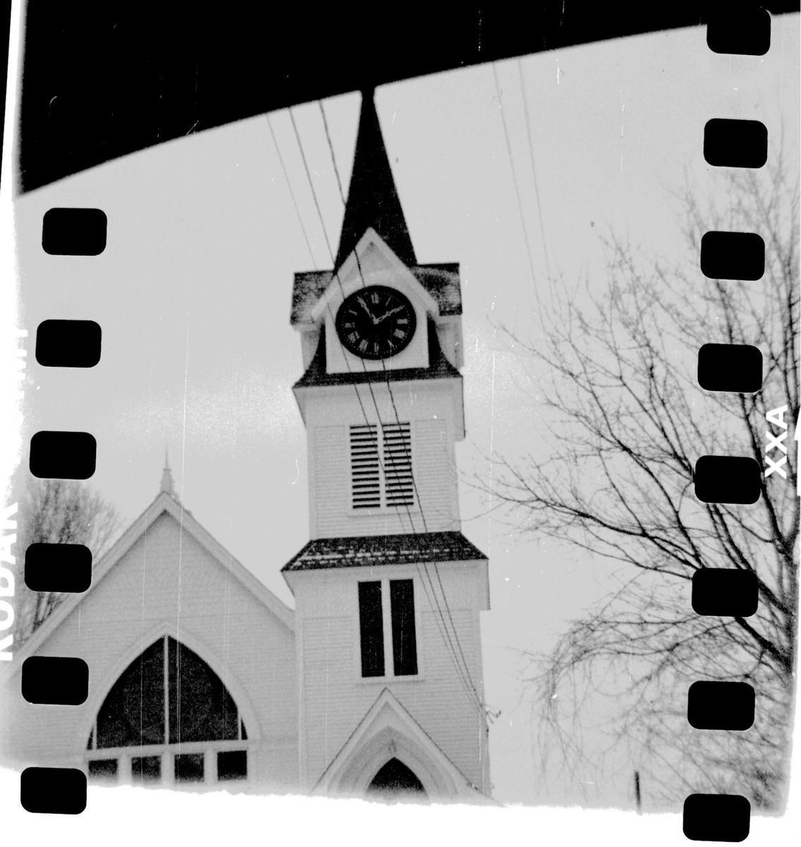 A white church with a tall steeple and clock tower stands next to a bare tree.