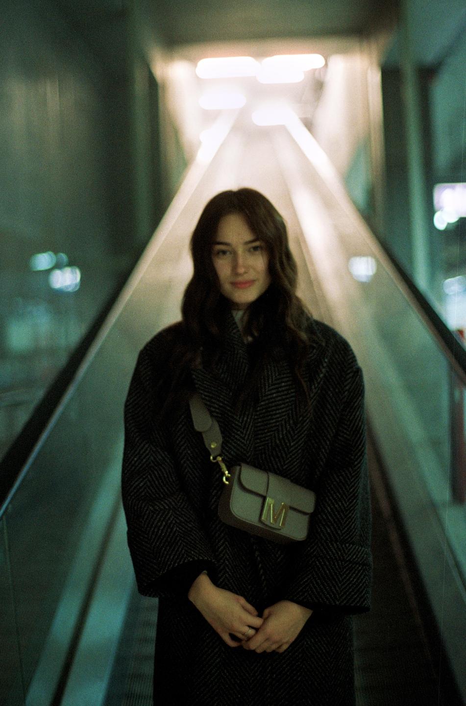 A woman stands on an escalator, looking forward with a slight smile.
