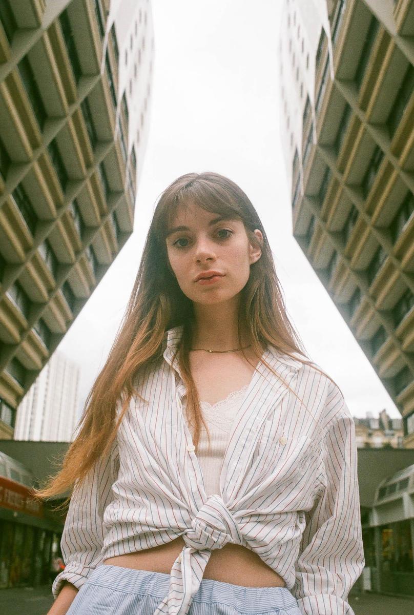 A woman with long hair and bangs stands between two tall buildings, looking directly at the viewer.