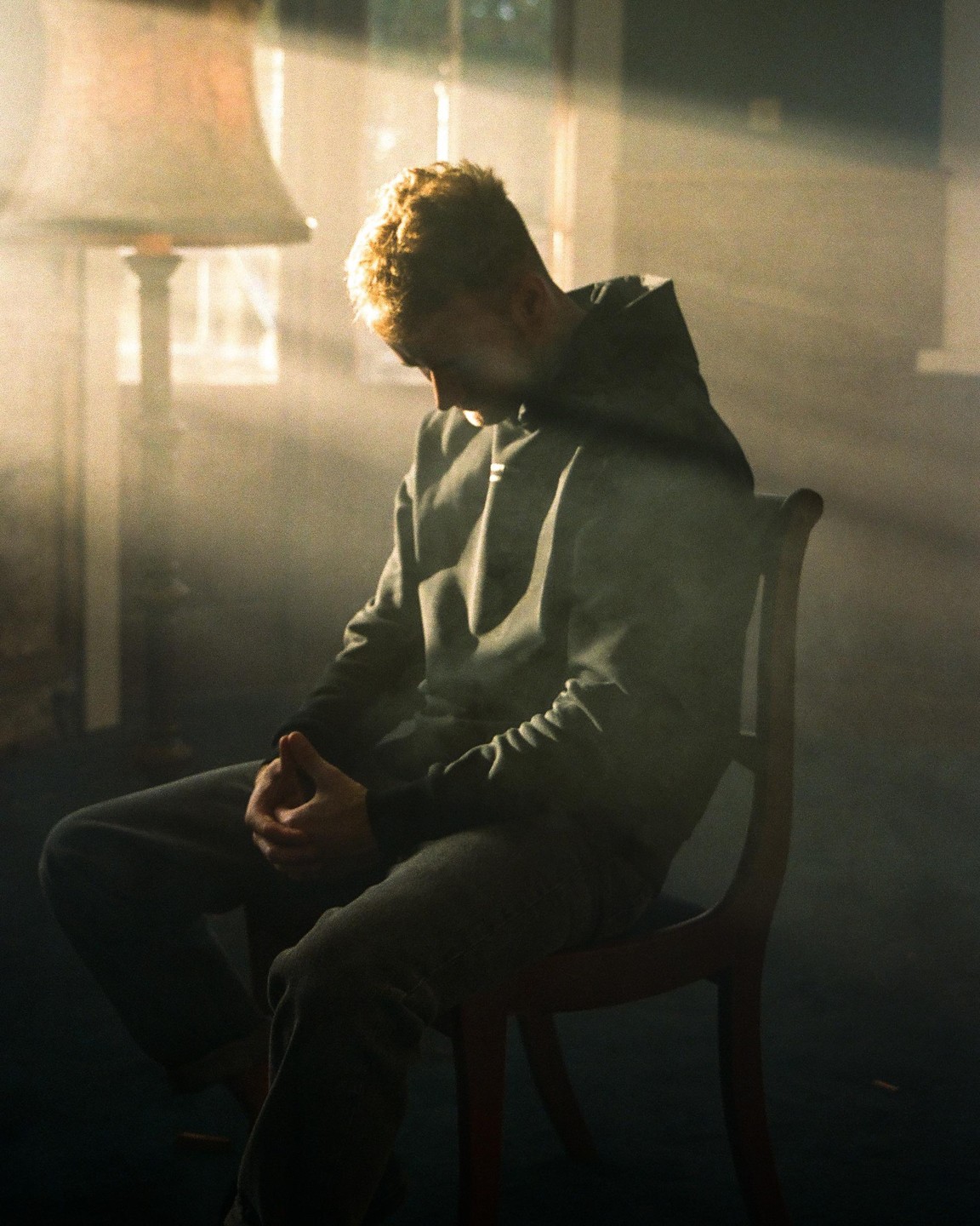 A man sits on a chair with his head bowed and hands clasped, illuminated by a shaft of light.