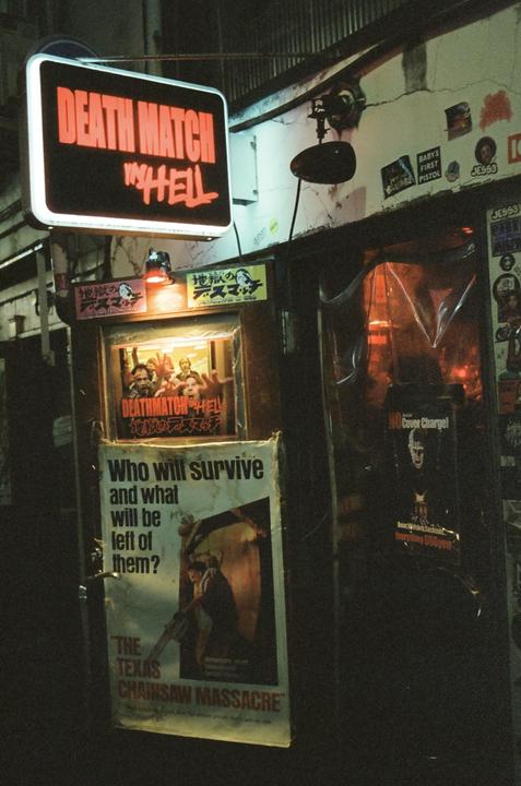 A neon sign for "Death Match in Hell" hangs above a poster for "The Texas Chainsaw Massacre" on a building at night.