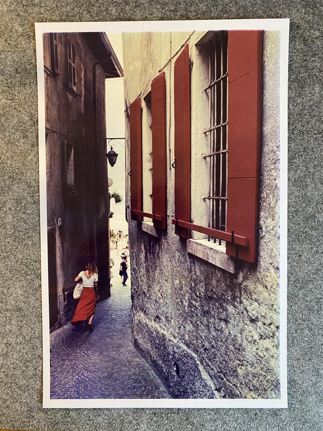 A woman in a red skirt walks down a narrow cobblestone alleyway between old buildings with red shutters.