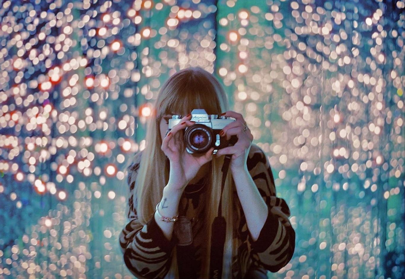 A woman holds a camera up to her face, obscuring it, against a background of bright, out-of-focus lights.