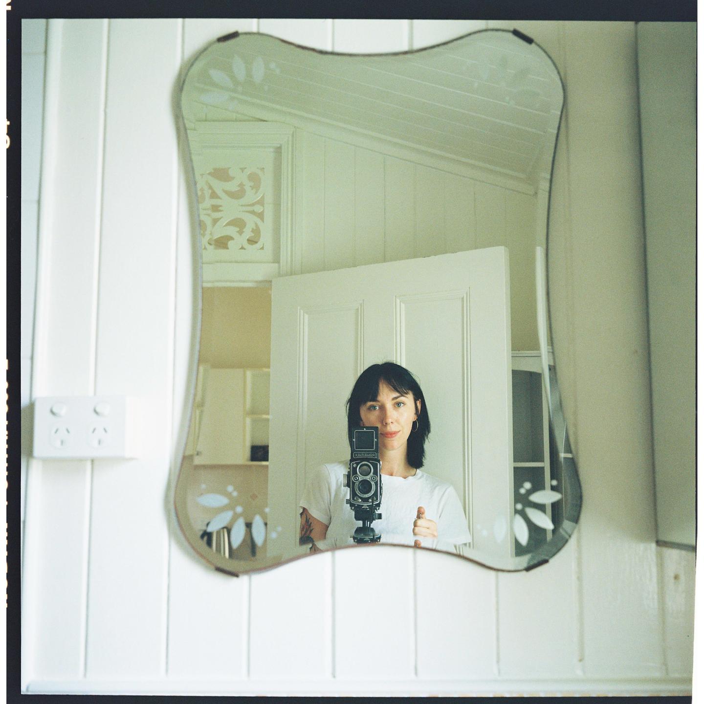 A woman with dark hair holds a camera and looks into a decorative mirror, capturing her reflection.