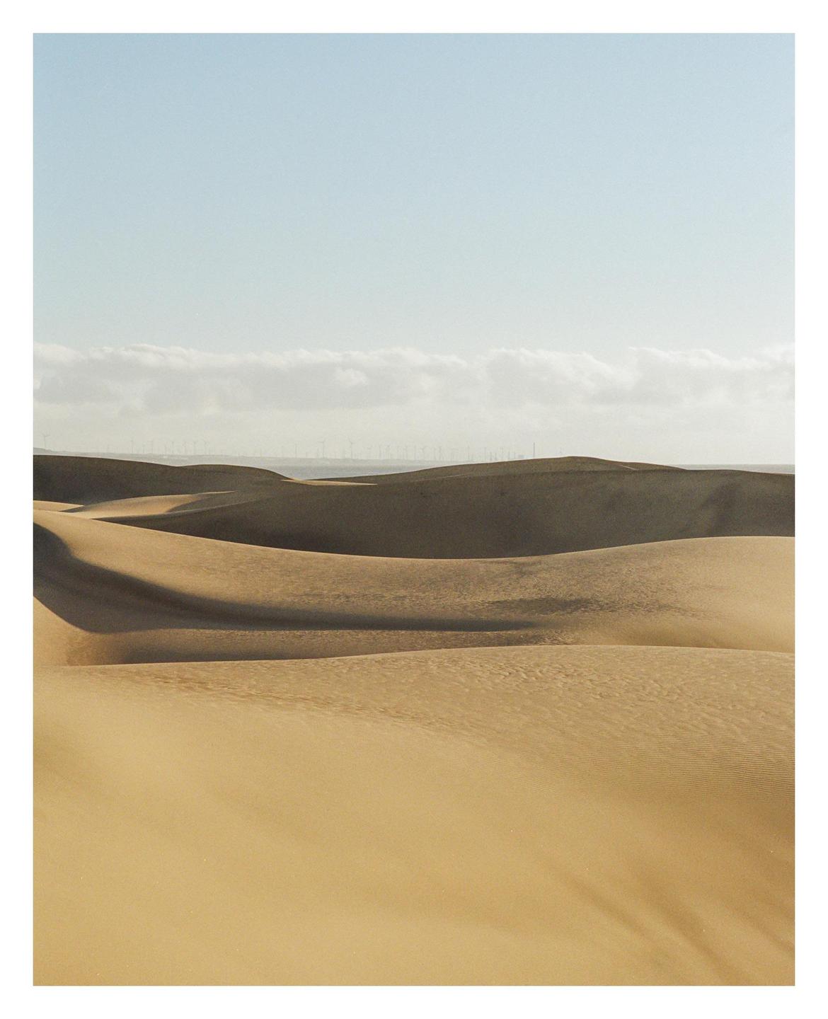 Rolling sand dunes stretch across the landscape under a clear sky with a distant line of wind turbines.