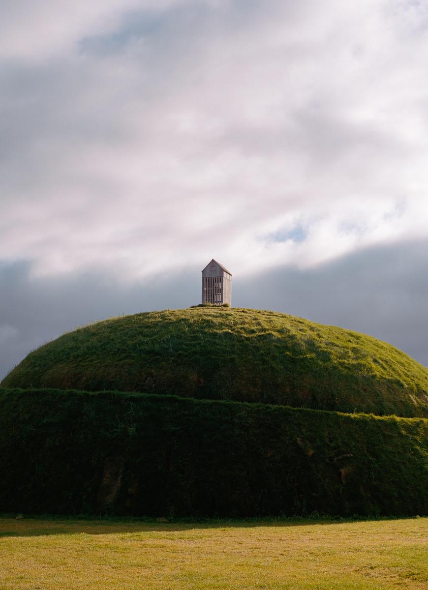 A small wooden structure sits atop a grassy hill under a cloudy sky.