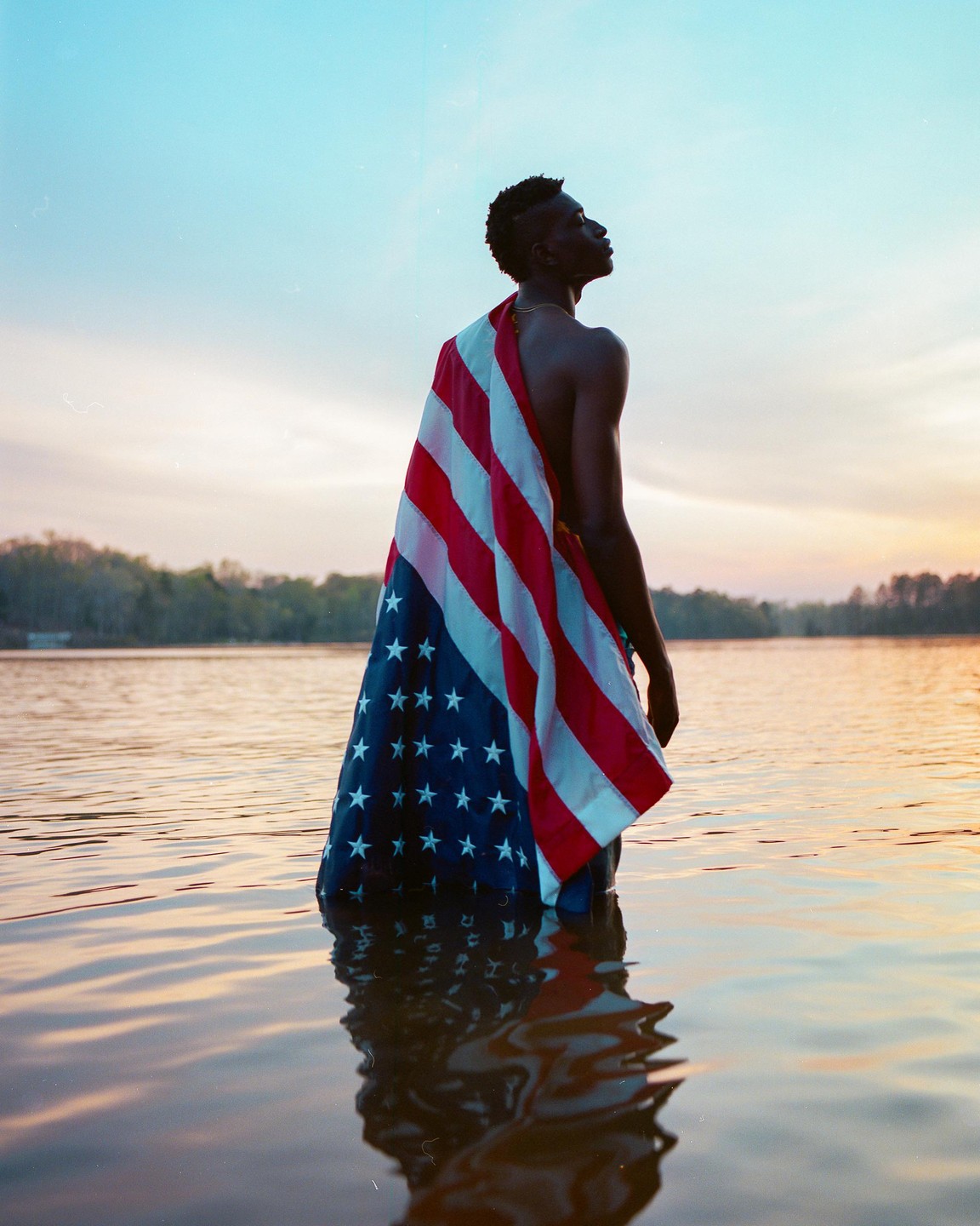 A man stands in water with an American flag draped over his shoulder at sunset.