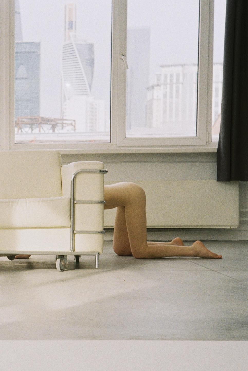 A person is kneeling on the floor behind a white sofa, with a city visible through a window in the background.