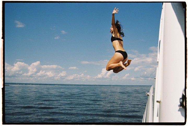 A woman in a black bikini jumps from a boat into the water under a blue sky with clouds.