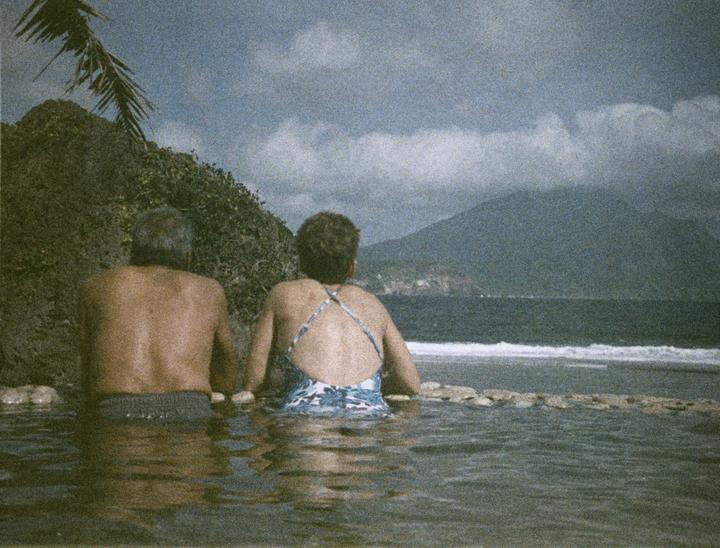 A couple sits in a pool of water looking out at the ocean and a distant mountain.