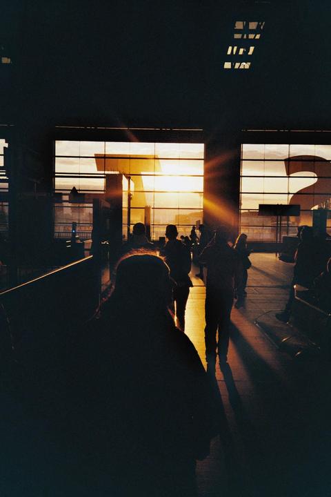 Silhouetted people stand in a building with large windows looking out onto a sunset.