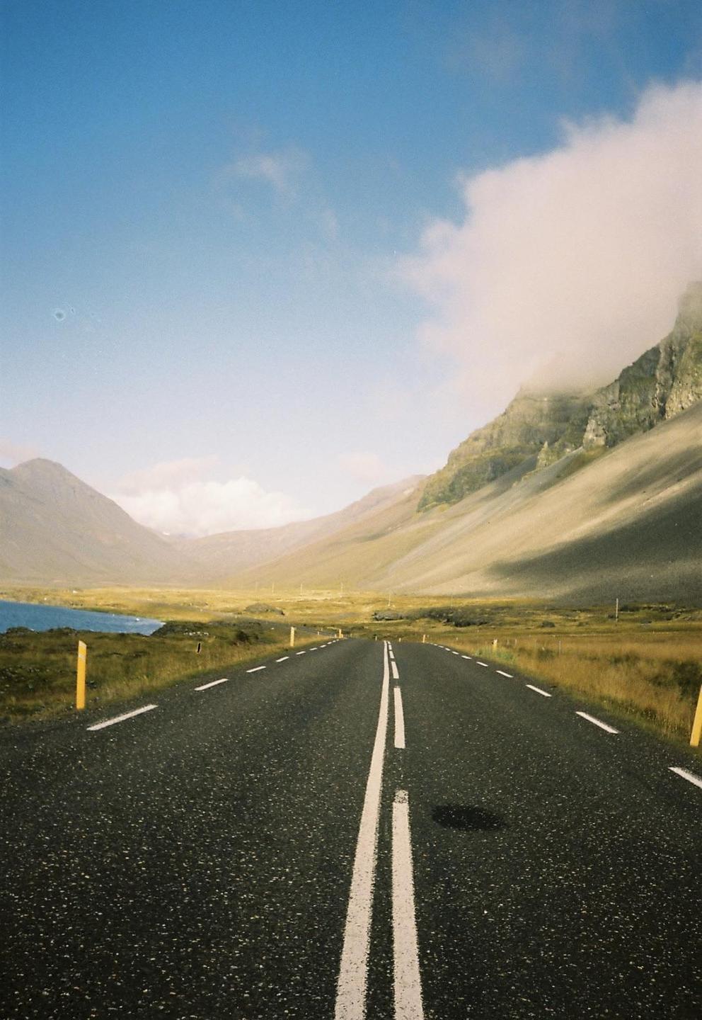A long road stretches into the distance, flanked by mountains and a body of water under a blue sky.
