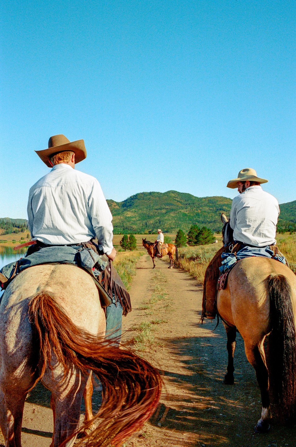 Three men on horseback ride along a dirt path next to a lake with mountains in the background.
