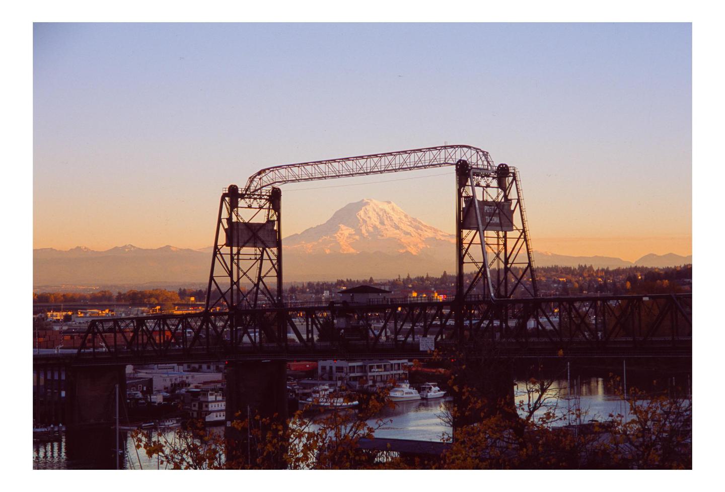 A snow-capped mountain is visible through the opening of a bridge over a body of water with boats.