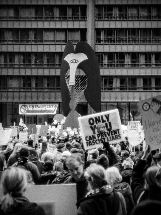 A large crowd of people are gathered in a street protest with signs and a large sculpture in the background.