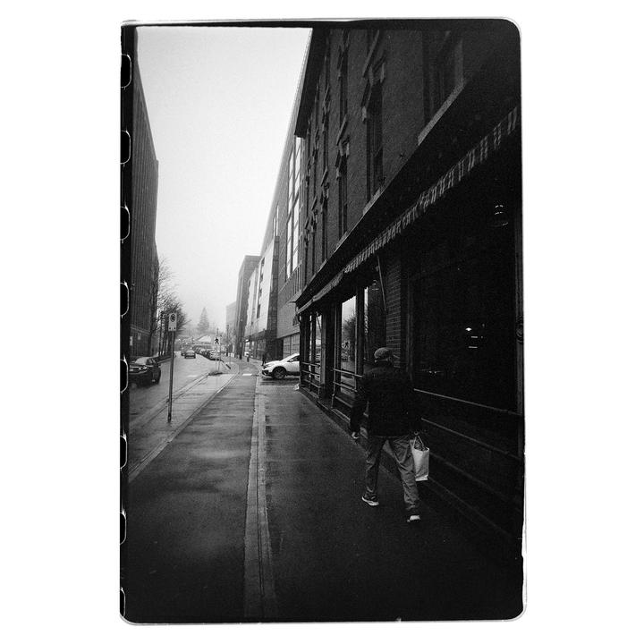 A man walks along a wet sidewalk next to a building with awnings in a hazy street scene.