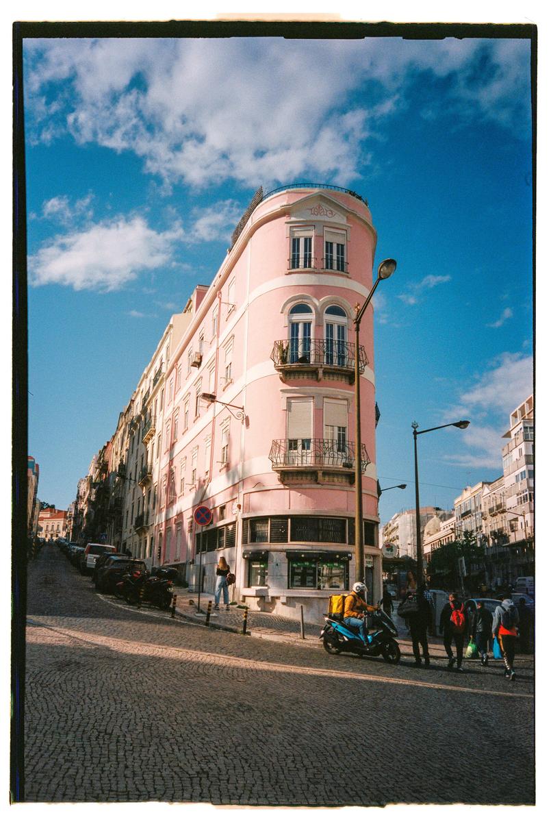 A pink building with balconies stands on a cobblestone street corner under a blue sky with clouds, with people and vehicles nearby.