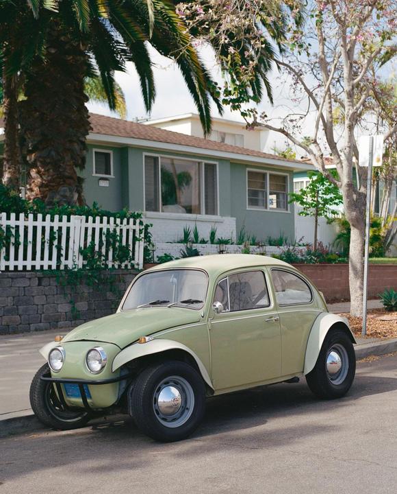 A light green Volkswagen Beetle is parked on a residential street in front of a house with a white picket fence and palm trees.