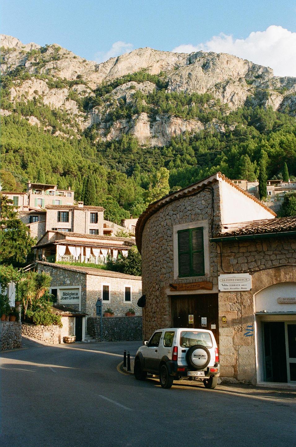A white Suzuki SUV is parked on a winding road in a village with stone buildings and a mountain in the background.