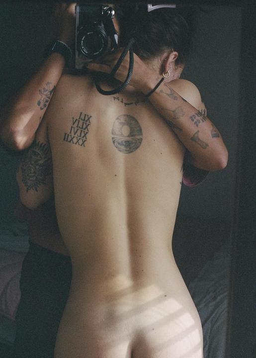 A person with tattoos on their back and arms is embraced by another person holding a camera.