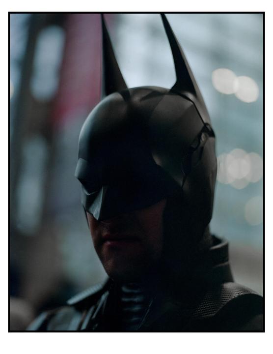 A man in a Batman costume is visible from the chest up, looking to the left.